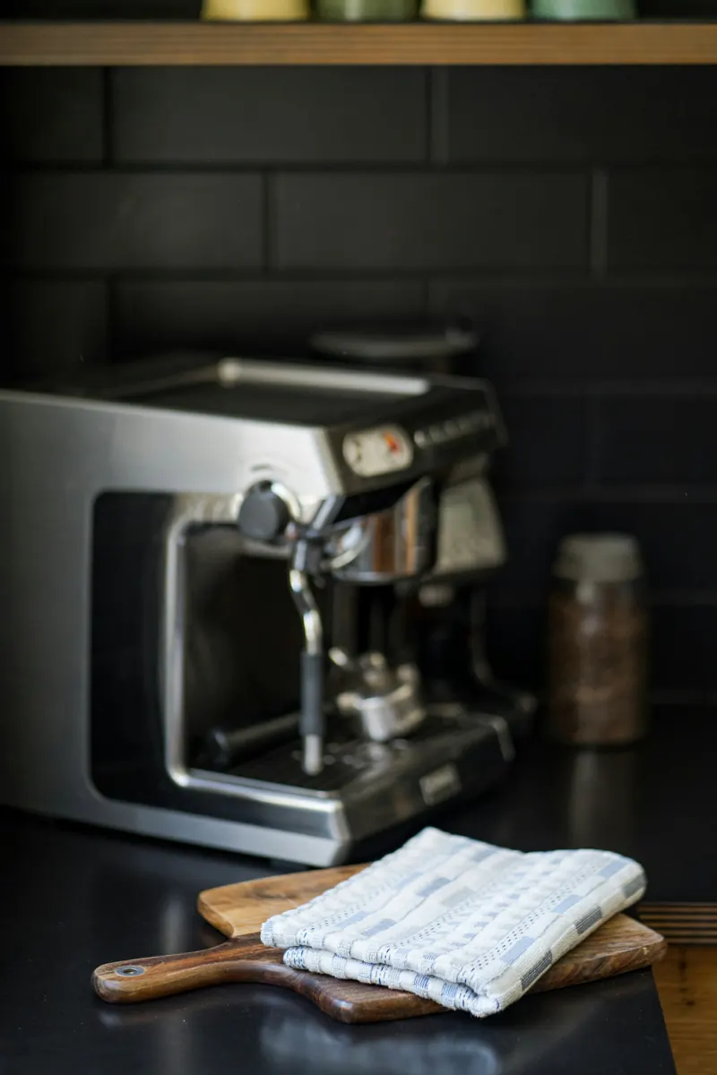 A stainless espresso machine on a black counter against dark tile, with a folded blue and white towel on a wooden board in the foreground and a jar of coffee beans behind.