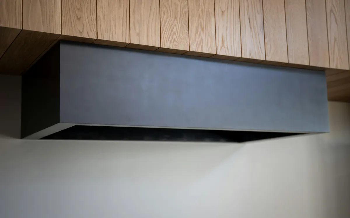 A low rectangular dark steel range hood mounted beneath vertical oak paneling, seen from below against a pale wall.