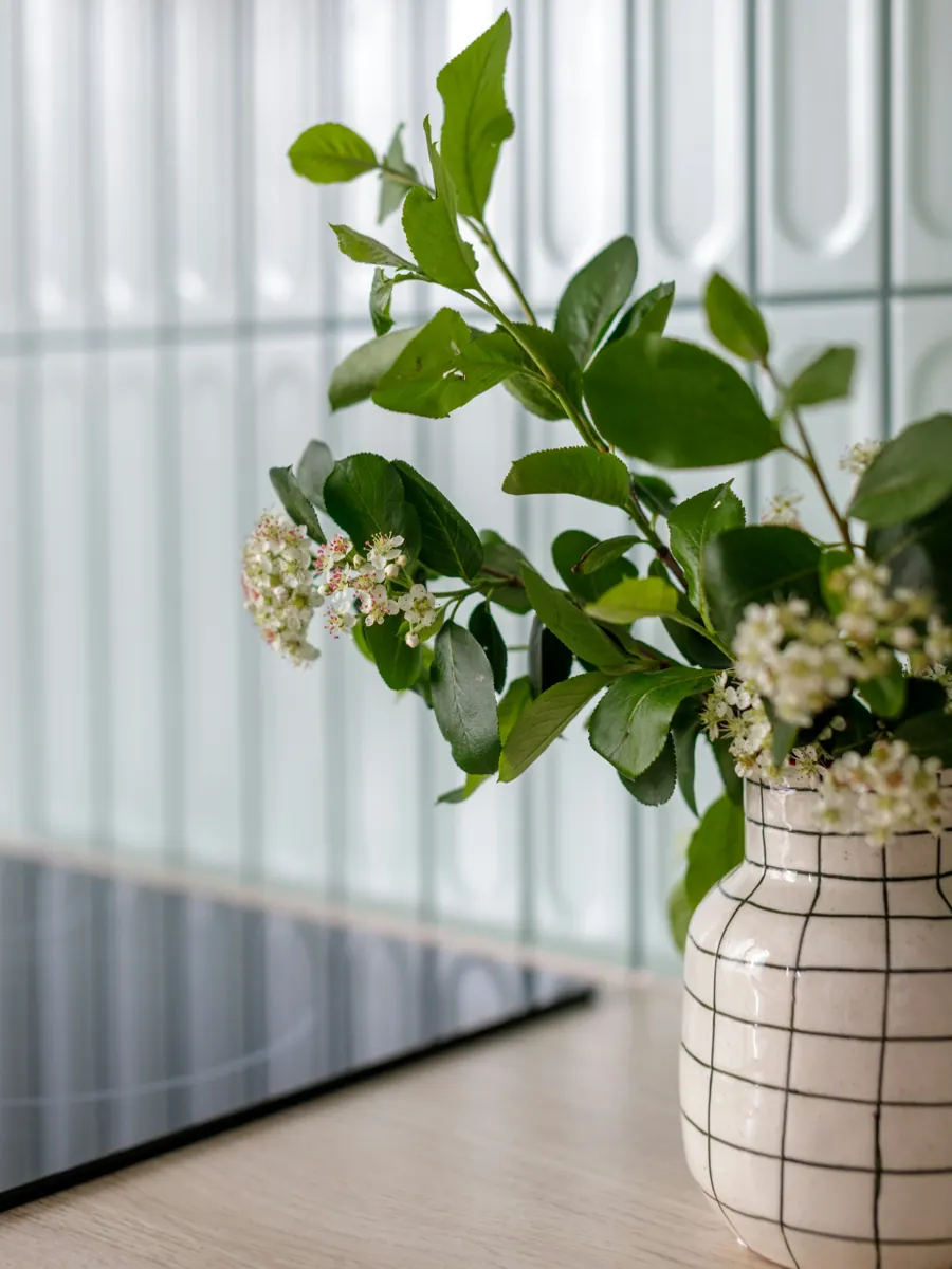 A ceramic vase with a grid pattern holding leafy branches with small white flowers, beside the edge of a black induction cooktop on a light oak counter, glazed tile behind.
