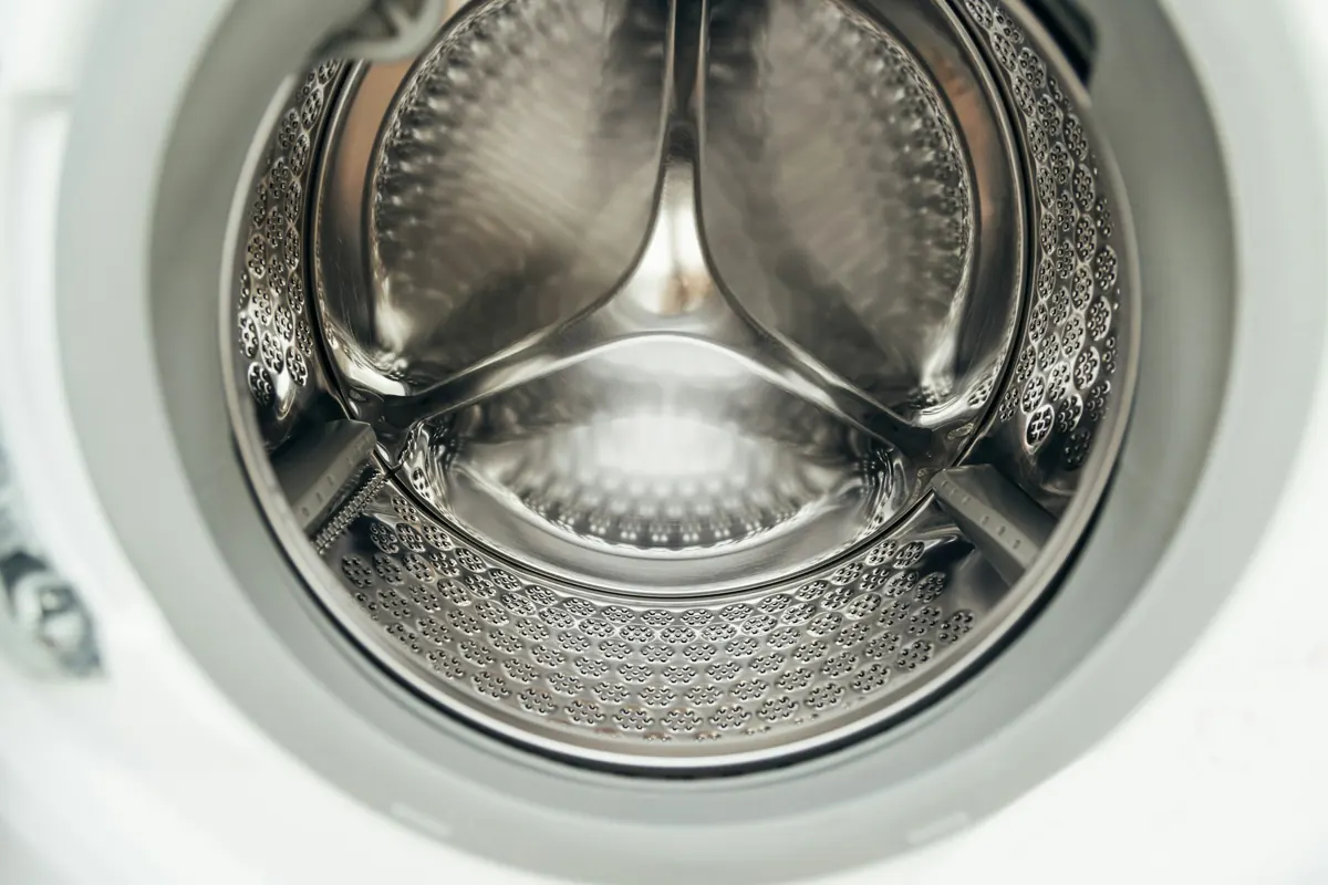 Looking straight into the empty stainless steel drum of a front-load washing machine, its perforated walls and three lifters catching soft light.