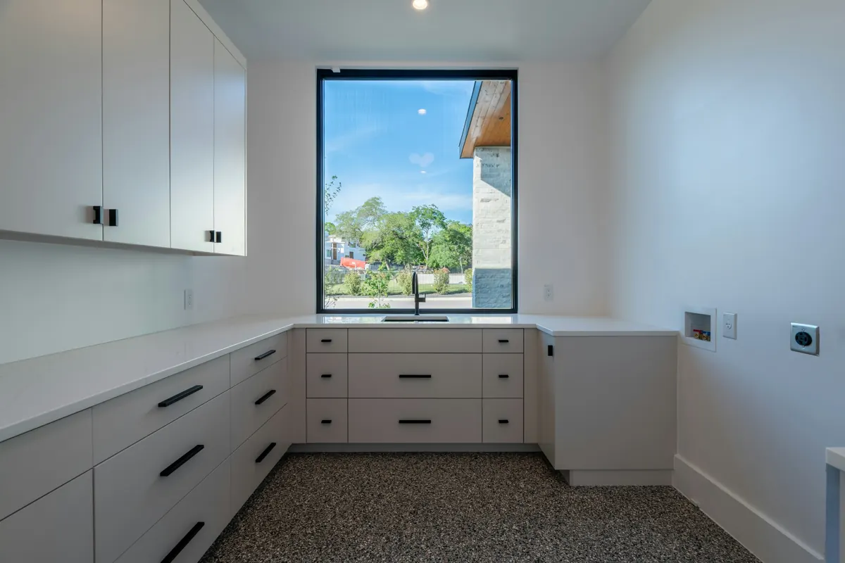 An empty laundry room with white flat-front cabinets, a white quartz counter running around a large window, a speckled floor, and a recessed washer box and a dryer power receptacle on the right-hand wall.