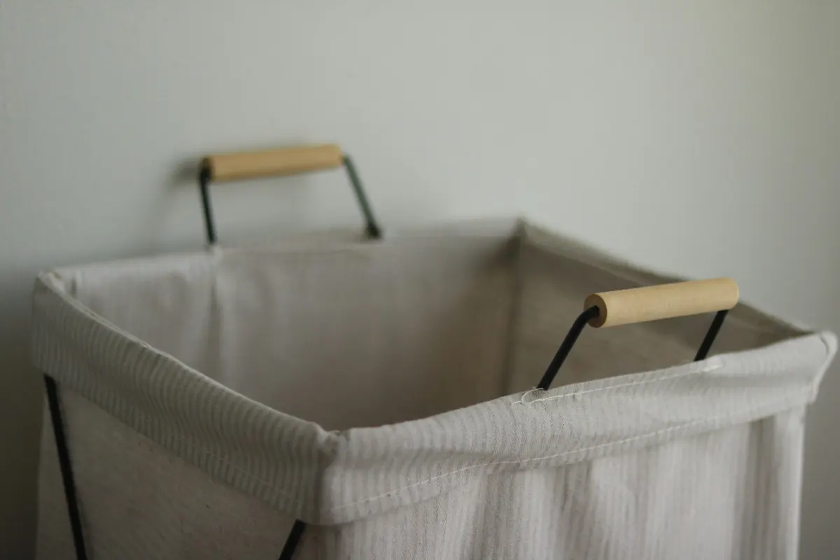 A soft linen laundry hamper with a black wire frame and wooden handles against a pale grey wall.