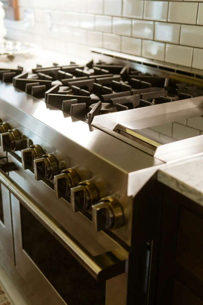 Close view of a stainless pro-style gas range with heavy cast-iron grates and a row of polished control knobs, white subway tile behind.