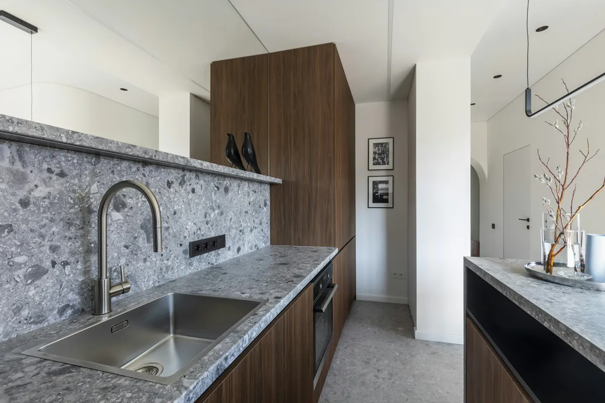 A galley kitchen with grey terrazzo counters and backsplash, a stainless single-bowl sink with a brushed gooseneck faucet, walnut base cabinets and a tall walnut cabinet, framed prints on the far wall.